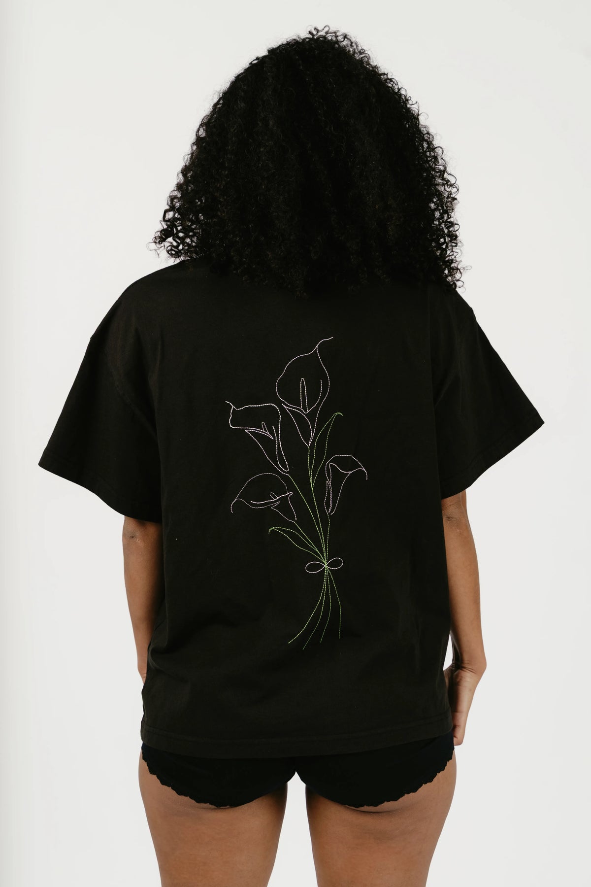 A person shown from the back wearing an oversized black T-shirt with a white line-art illustration of calla lilies on the back, paired with black shorts against a plain light background.