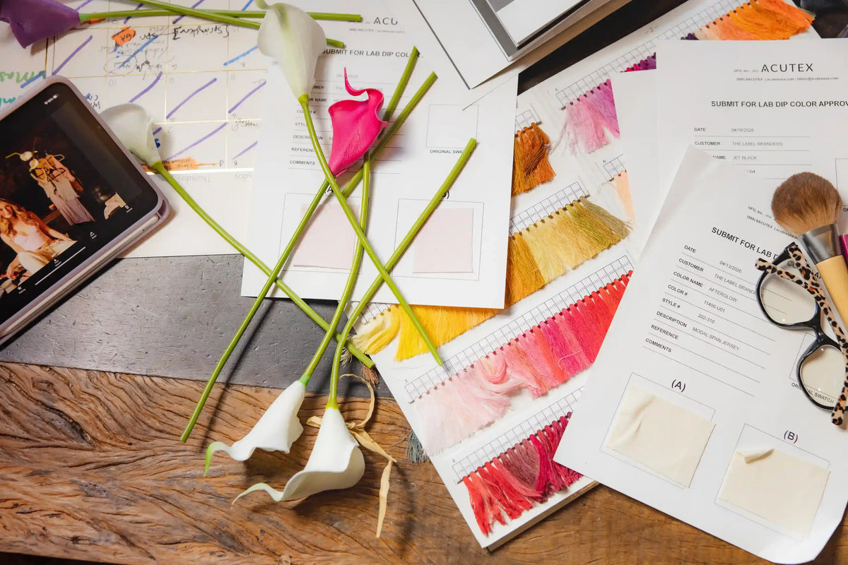 Callah behind-the-scenes flat lay of fabric dye lab submissions, thread color 
swatches, and design notes spread across a wooden table with fresh calla lilies