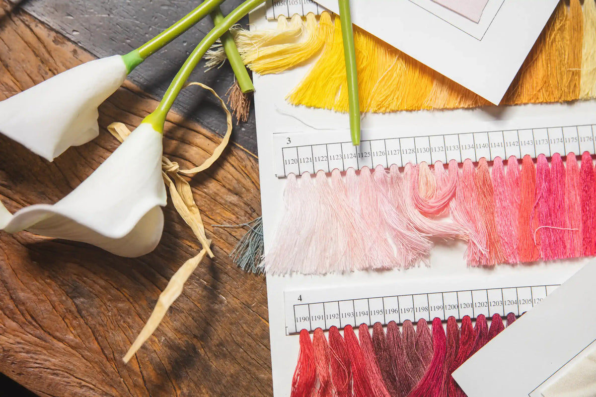 Callah behind-the-scenes flat lay of thread color swatches in blush, pink, 
and yellow tones alongside white calla lilies on a wooden table during the 
design process